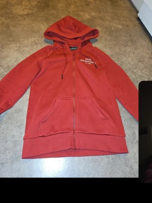 Röd hoodie från Peak Performance - Snygg röd hoodie från Peak Performance med broderad logga på bröstet. Tröjan har dragkedja, huva med snören och stora framfickor. Perfekt för chill dagar och har en klassisk passform. Materialet är mjukt och bekvämt, troligen bomullsblandning.