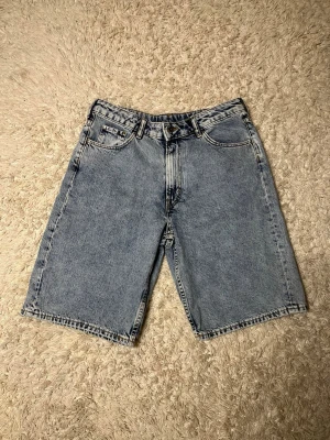 Blå denim shorts med klassisk passform - Säljer ett par blå denim shorts med klassisk femficksdesign och normal passform. Shortsens längd går till knäna och de har en ljus tvätt med dragkedja och knapp framtill. Perfekta för sommarens chill dagar.