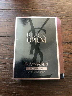 Yves Saint Laurent Black Opium EdP. 1,2 ml - Black Opium Eau de Parfum från Yves Saint Laurent är en ikonisk parfym med en elegant svart och roséguldig förpackning. Doften är känd för sin djupa, sensuella karaktär och flaskan har en modern, lyxig design med YSL-loggan i bakgrunden.