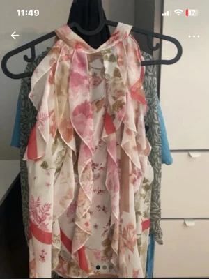 Blommig volangblus med hög krage - Säljer en romantisk blus med blommigt mönster i rosa, grönt och rött. Blusen har volangdetaljer framtill och en hög krage som knäpps i nacken. Den är ärmlös och gjord i ett lätt, transparent material som känns luftigt och somrigt.