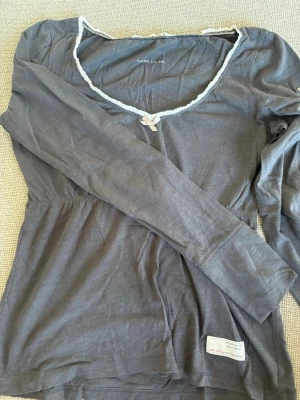 Grå långärmad topp Odd Molly - Supersöt grå långärmad topp från Odd Molly med vit spetskantad v-ringning och liten rosett framtill. På ena ärmen finns en broderad detalj med fågel och Odd Molly-logga. Tillverkad i mjuk bomull, perfekt för en chill och snygg look.