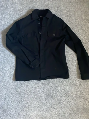 Svart overshirt från Gabba - Stilren svart overshirt från Gabba med klassisk krage och knäppning framtill. Två bröstfickor med knapp, perfekt att bära som lager över t-shirt eller hoodie.