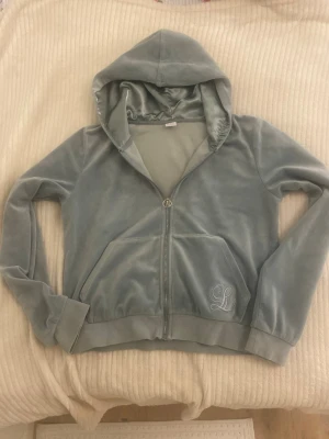 Ljusblå velour hoodie med dragkedja - Hej! Jag säljer en ljusblå hoodie i mjuk velour med dragkedja och huva. Tröjan har två stora fickor framtill och ett broderat L på ena fickan. Skulle gärna rekommendera att ”styla” den med ett vitt linne under och kanske ett guldhalsband! Pris kan diskuteras, men skulle säga cirka 100-150kr.