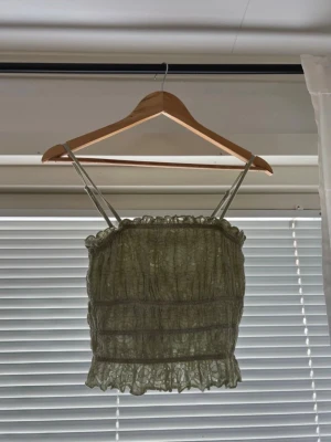 Grön linnetopp med volangdetaljer - Säljer en ljusgrön linnetopp med smala axelband och volangdetaljer längs både över- och nederkant. Toppen har ett rynkat och transparent material som ger en luftig känsla. Perfekt för varma dagar och snygg att styla med jeans eller kjol.