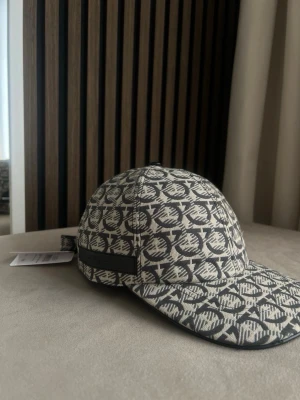 Beige keps med svart mönster från ferragamo - Säljer en beige keps från ferragamo med svart grafiskt mönster över hela kepsen. Klassisk form med böjd skärm och justerbar rem baktill. Materialet är textil och kepsen har en lyxig känsla med tydlig logotypdesign.