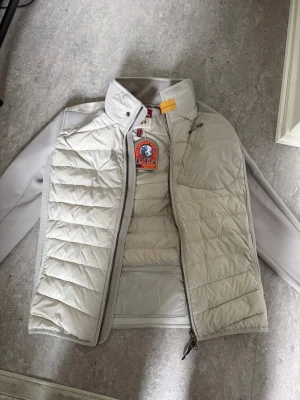 Ljusgrå pufferjacka från Parajumpers - Säljer en ljusgrå pufferjacka från Parajumpers med quiltad front och släta paneler. Jackan har hög krage, dragkedja och en ikonisk patch på ärmen. Materialet är en kombination av syntet och vaddering, perfekt för kalla dagar.