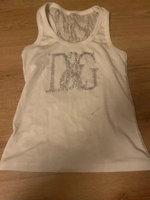 linne D&G med spets - Säljer en vit linnetopp från D&G med glittriga stenar som bildar logotypen på bröstet. Toppen har snygg spetsdetalj i ryggen som ger en extra touch. Perfekt för dig som gillar stilrena och trendiga plagg.