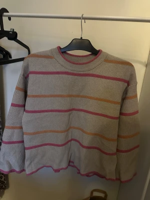 Beige stickad tröja med ränder - Säljer en beige stickad tröja med ribbad struktur och färgglada ränder i rosa och orange. Tröjan har rund halsringning och avslut med vågig kant. Perfekt för dig som gillar en avslappnad och trendig stil.