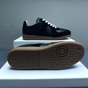 Svarta Maison Margiela Gats - Stilrena svarta Maison Margiela Gats med vit insida och vita skosnören. Skorna har en klassisk låg profil och en kontrasterande ljusbrun gummisula. Materialet är en mix av läder och mocka, vilket ger en snygg och modern look.