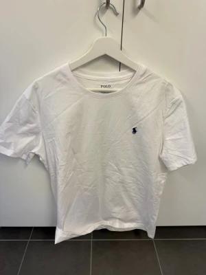 Vit t-shirt från Polo Ralph Lauren - Klassisk vit t-shirt från Polo Ralph Lauren med rund halsringning och korta ärmar. T-shirten har den ikoniska lilla marinblå logotypen broderad på bröstet. Tillverkad i mjuk bomull som känns skön mot huden. Perfekt basplagg till garderoben.