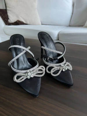 Svarta pumps med strass  - Eleganta svarta pumps New Yorker med spetsig tå och höga klackar. Skorna har glittrande strassband som korsar foten och en dekorativ rosett med strass och kristaller framtill. Endast använda en gång. Ca 10 cm höga