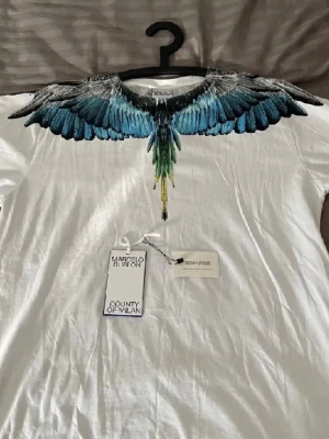Vit t-shirt med blått fågelmotiv - Säljer en vit t-shirt från Marcelo Burlon County of Milan med ett coolt fågelmotiv i blått, svart och grönt runt halsen. T-shirten har korta ärmar och är gjord i mjuk bomull. Perfekt för dig som vill sticka ut med en unik design. Den är i väldigt bra skick och nogrant omhändetagen.
