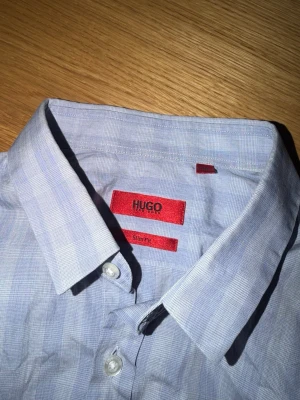 Blårandig skjorta från Hugo Boss - Säljer en stilren blårandig skjorta från Hugo Boss med slim fit-passform. Skjortan har klassisk krage, vita knappar och lång ärm. Perfekt för dig som vill ha en snygg och modern look. Materialet är bomull och mönstret är diskret randigt.