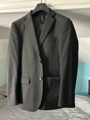 Kostym Dressmann Svart Slim fit - Enkel svart kostym från Dressman i slim fit modell. Storlek 46 på kavajen och byxorna är 30/30. Materialet är en ullblandning. Kostymen är använd nästan bara en gång och det var på skolavslutningen i nian (2023). Tror jag var typ 170 lång då. 