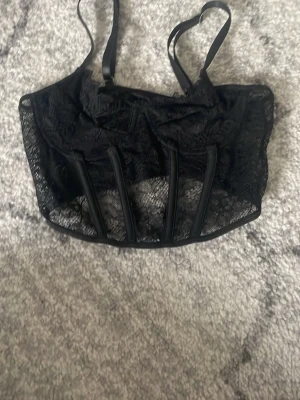 Svart spetskorsett med stålstavar - Snygg svart korsett med justerbara axelband och dekorativ spets. Korsetten har genomskinliga meshpartier och flera stålstavar framtill som ger en cool och strukturerad look. Perfekt för att lyfta din outfit med en edgy touch.