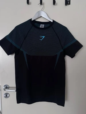 Gymshark Onyx v5 - Snygg svart tränings-tshirt från Gymshark med korta ärmar och blått mönster över bröst och axlar. Gymshark-loggan i blått framtill. Tillverkad i ett lätt och stretchigt funktionsmaterial som andas, perfekt för gymmet eller löpning.