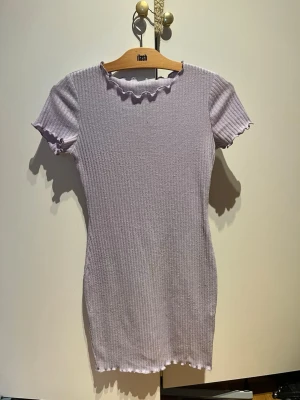 Ljuslila ribbad kortklänning - Säljer en ljuslila ribbad kortklänning med volangdetaljer vid hals, ärmslut och nederkant. Klänningen har en slim passform och är kortärmad, perfekt för dig som gillar en enkel men söt stil.