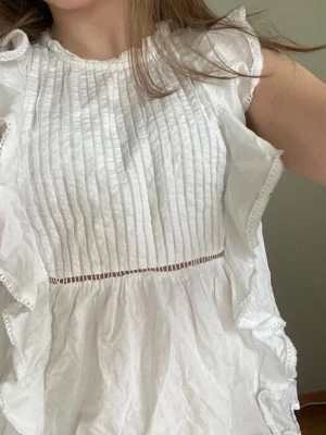 Vit blus med volanger från BikBok - Supersöt vit blus från BikBok med volangdetaljer längs sidorna och smockad framsida. Blusen har rund halsringning, tunna spetskanter och är tillverkad i ett lätt, krispigt tyg. Perfekt för en fräsch och trendig look.