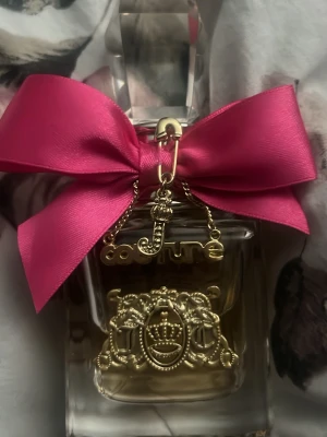 Äkta Juicy Couture parfym med rosa rosett - Lyxig parfymflaska från Juicy Couture med en stor, glittrig rosa rosett och guldfärgade detaljer. Flaskan är genomskinlig med ett elegant emblem framtill och en dekorativ kristallformad kork. Perfekt för dig som gillar glam och snygg design.