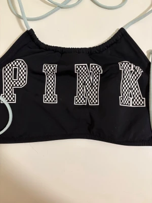 Svart bikiniöverdel från PINK - Snygg svart bikiniöverdel från PINK med vita snören och stora bokstäver framtill. Bokstäverna har ett coolt mönster i vitt och svart. Perfekt för stranden eller poolen!