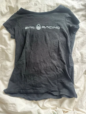 Svart Sail Racing t-shirt - Svart t-shirt från Sail Racing med vit logotyp på bröstet och liten logga i nacken. Klassisk passform och rund hals, perfekt för dig som gillar sportiga och stilrena plagg. Materialet känns mjukt och lätt, vilket gör den skön att bära hela dagen. Den har även hål där framme som är slitning