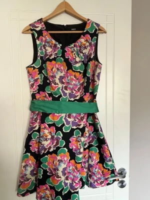 Blommig klänning från Oasis - Färgglad blommig klänning från Oasis med svart bas, stora blommor i lila, orange, rosa och grönt samt ett grönt midjeband. Ärmlös modell med rund hals och lätt utställd kjol. Perfekt för dig som gillar statement-plagg!