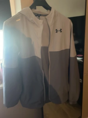 helt ny, Grå vindjacka från Under Armour - men dragkedjan e sönder men de kostar bara typ 100 kr laga den Snygg tvåtonad grå vindjacka från Under Armour med huva och dragkedja framtill. Jackan har logga på bröstet och elastiska muddar vid ärmsluten. Perfekt för blåsiga dagar och sportiga outfits.