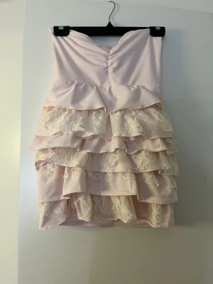 Ljusrosa axelbandslös volangtopp - Söt axelbandslös tubtopp i ljusrosa med flera lager volanger och spetsdetaljer. Toppen har en bandeau-form och romantisk känsla med blommig spets och omlottvolanger. Perfekt för dig som vill ha en feminin och trendig look. Denna toppen har jag gjort själv. Tyget är stretchigt och vid ryggen finns det resår för att inte riskera att toppen faller ner. Storleken är Xs/S