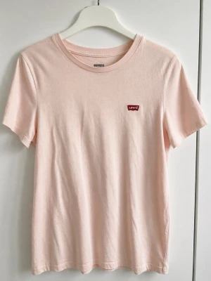 Ljusrosa Levi's t-shirt i bomull - Säljer en ljusrosa t-shirt från Levi's med klassisk rund hals och korta ärmar. T-shirten har en liten röd Levi's-logga på bröstet och är gjord i mjuk bomull. Perfekt basic-plagg för en clean och enkel stil.