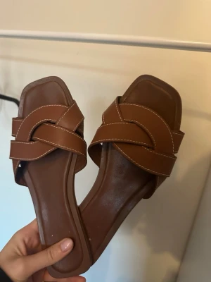 Bruna slip-in sandaler med flätad rem - Snygga bruna slip-in sandaler med flätad remdesign och vita kontrastsömmar. Sandalerna har fyrkantig tå och är tillverkade i konstläder. Perfekta för en trendig och avslappnad look.