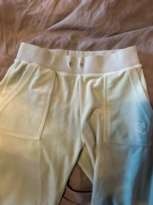 Ljusgröna mjukisbyxor från Juicy Couture - Snygga ljusgröna mjukisbyxor från Juicy Couture i mjuk velour. Byxorna har bred resår i midjan, stora fickor fram och bakfickor med lock och tryckknappar. Juicy Couture-logga broderad på ena fickan framtill. Perfekta för chill dagar. SAKNAR SNÖRE!