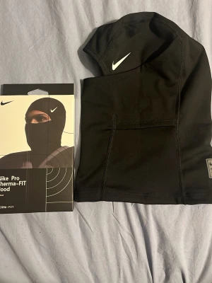 Nike Pro Therma-FIT balaclava - Nike Pro Therma-FIT balaclava i svart, tillverkad i ett mjukt och stretchigt material som håller värmen. Snygg vit Nike-logga på sidan. Perfekt för träning utomhus eller kalla dagar. Säljes med originalförpackning.