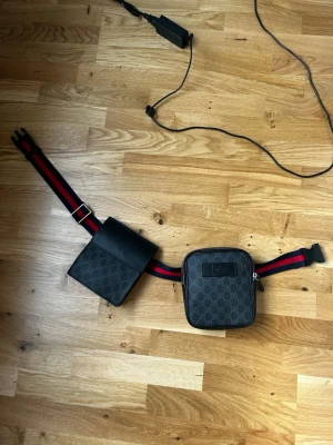 Gucci GG Supreme bälte-väska svart - Snygg bälte-väska från Gucci i svart GG Supreme canvas med två fack. Väskan har justerbart band i rött och blått, samt detaljer i svart läder. Perfekt för att bära det viktigaste och ger en trendig streetwear-look.