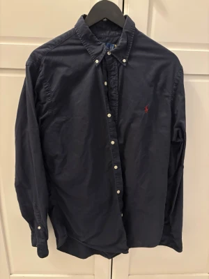 Mörkblå skjorta från Polo Ralph Lauren - Säljer en klassisk mörkblå skjorta från Polo Ralph Lauren med vit knappstängning och broderad logga i rött på bröstet. Skjortan har lång ärm och button-down krage, perfekt för en stilren look.