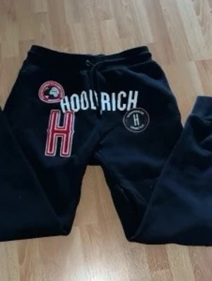 Svarta Hoodrich mjukisbyxor - Säljer ett par svarta mjukisbyxor från Hoodrich med stora tryckta loggor och text på framsidan. Byxorna har dragsko i midjan och muddar vid bensluten. Perfekta för en avslappnad och streetinspirerad stil.