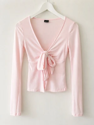 Ljusrosa topp med knyt framtill - Supersöt ljusrosa topp med långa ärmar och djup V-ringning. Toppen har en snygg knytning framtill som ger en feminin touch. Tillverkad i mjukt och stretchigt material som sitter skönt på kroppen. Perfekt att styla med jeans eller kjol.