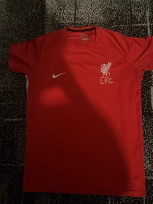 Liverpool FC röd Nike matchtröja - Säljer en röd Liverpool FC matchtröja från Nike med klubbens logga och Nike-swoosh i vitt på bröstet. Tröjan har korta ärmar och vita detaljer längs sidorna. Perfekt för dig som älskar fotboll och vill stötta LFC på match eller träning.