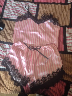 Rosa randigt satinset med spets - Sött tvådelat set i rosa och vitt satin med vertikala ränder. Både linne och shorts har tunna axelband och dekoreras med svart spets vid kanterna. Shortsen har en svart rosett i midjan. Perfekt för dig som gillar en feminin och lyxig känsla.