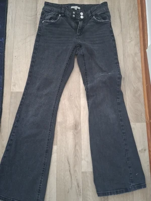 Svarta bootcut jeans från Denim Day - Säljer ett par svarta bootcut jeans från Denim Day i storlek 164. Jeansen har klassiska femficksdetaljer, silverfärgade knappar och är tillverkade i denim. Perfekt för dig som gillar en lite vidare passform nertill.