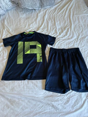 Blå sportset med neontryck - Säljer ett blå sportset med t-shirt och shorts. T-shirten har ett stort neonfärgat tryck med siffran 19 och texten 'Soccer Pro' på framsidan. Båda plaggen är gjorda i ett lätt och snabbtorkande material, perfekt för träning eller fotboll.