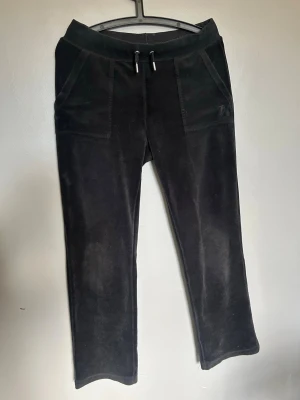 Svarta velourbyxor Juicy Couture - Mjukisbyxor i svart velour från Juicy Couture med bred resår i midjan och dragsko. Byxorna har två fickor fram och två bakfickor med knappar. Klassisk logga broderad på ena fickan framtill. Perfekta för en chill och avslappnad stil.