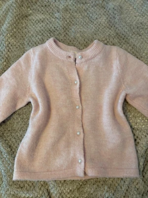 Ljusrosa stickad kofta med pärlknappar - Söt ljusrosa stickad kofta med pärlknappar framtill och rund halsringning. Koftan har korta ärmar och är gjord i ett mjukt material som känns skönt mot huden. Perfekt att slänga över en topp för en chill look.