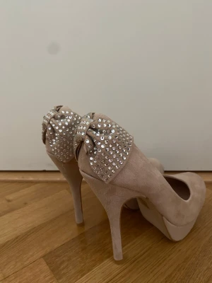 Beige pumps med rosett och strass - Snygga Steve Madden pumps med höga klackar och platåsula. Baktill pryds skorna av stora rosetter täckta med glittrande strass, vilket ger en extra glamorös touch. Materialet ser ut att vara mockaimitation. Perfekta för dig som vill sticka ut.