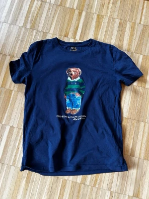 Polo Bear t-shirt från Ralph Lauren - Snygg marinblå t-shirt från Ralph Lauren med den ikoniska Polo Bear-trycket på bröstet. Klassisk passform och rund halsringning. Tillverkad i mjuk bomull som känns skön mot huden. Perfekt för dig som gillar stilrena plagg med en twist. Storlek S