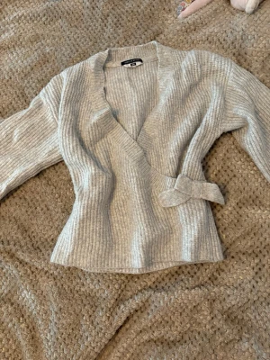 Ljusgrå omlottkofta  - Säljer en ljusgrå ribbstickad omlottkofta med knytband i sidan. Koftan har en djup v-ringning och långa ärmar, perfekt för lager på lager. Materialet är mjukt och stretchigt för en bekväm passform.