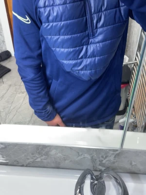 Blå hoodie från Nike med quiltad front - Snygg blå hoodie från Nike med quiltad, vadderad frontpanel och halv dragkedja. Tröjan har långa ärmar och en sportig look med Nike-logga på ärmen. Perfekt för dig som gillar streetwear och vill ha något bekvämt och stilrent.