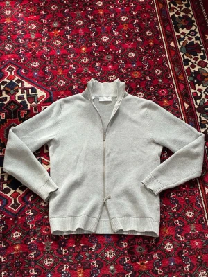 Ljusgrå ziptröja från Selected Homme - Ljusgrå stickad ziptröja från Selected Homme med hög krage och ribbade muddar. Tröjan har en diskret struktur i materialet och dragkedja framtill. Perfekt för lager på lager och enkel att matcha med olika outfits.