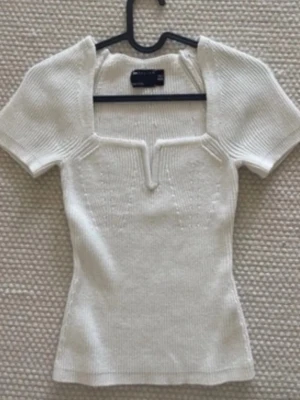 Vit ribbad topp  - Snygg vit ribbad topp  med korta ärmar och unik v-ringad detalj framtill. Toppen har en figurnära passform och är tillverkad i mjukt stickat material som känns skönt mot huden. Perfekt för att skapa en stilren look.