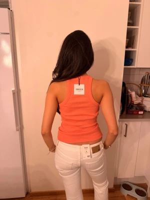 Koralröd ribbad tanktop från Edited - Säljer en koralröd ribbad tanktop från Edited med racerback och tight passform. Toppen är gjord i ett stretchigt material som sitter snyggt och bekvämt. Perfekt för att skapa en cool och avslappnad look.
