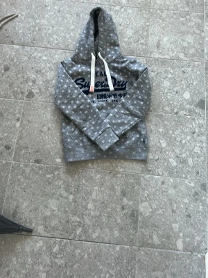 Grå hoodie Superdry med stjärnor - Grå hoodie från Superdry med vita stjärnor över hela tröjan och tryckt logga i marinblått framtill. Klassisk huva med vita snören och mjukt material som känns skönt mot huden. Perfekt för dig som gillar en avslappnad och cool stil.